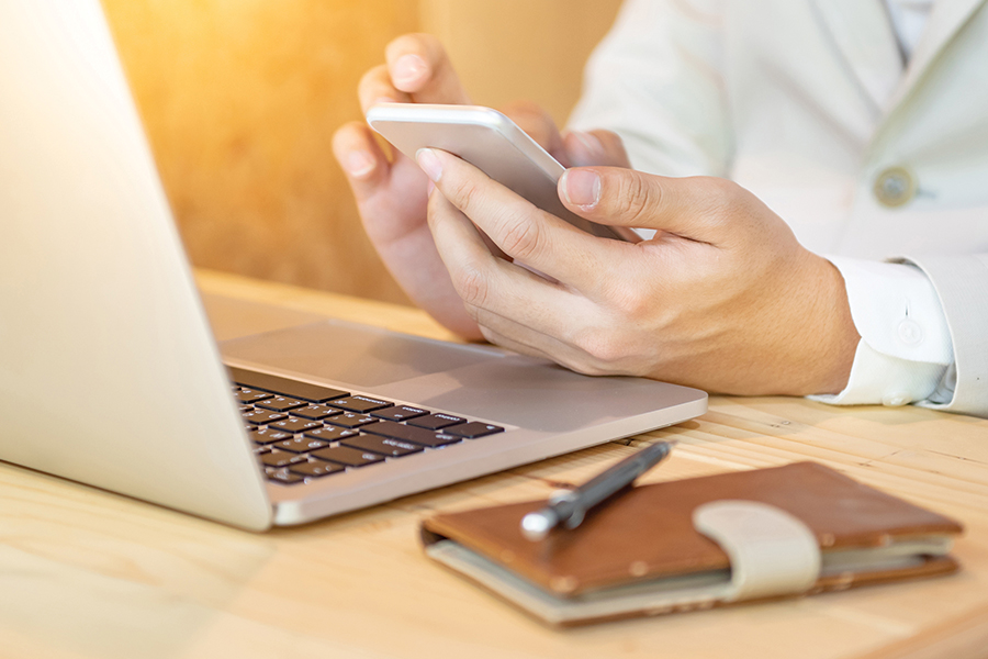 A person at a laptop scrolling on their phone.