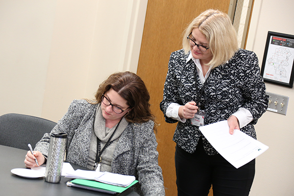 a woman holding a sheet of paper looking down at another woman writing in a notebook